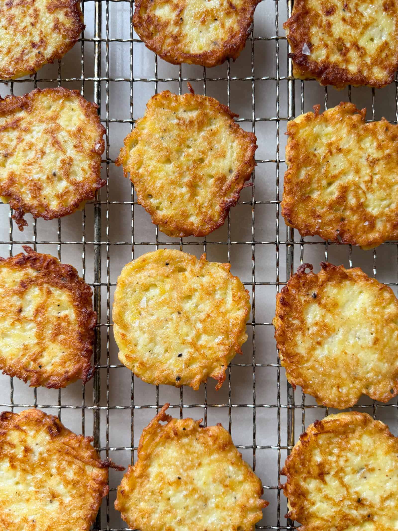 Golden brown potato latkes arranged in neat rows on a wire cooling rack, seen from above. Their crispy edges and textured surfaces showcase the irresistible appeal of a classic Potato Pancakes Recipe.