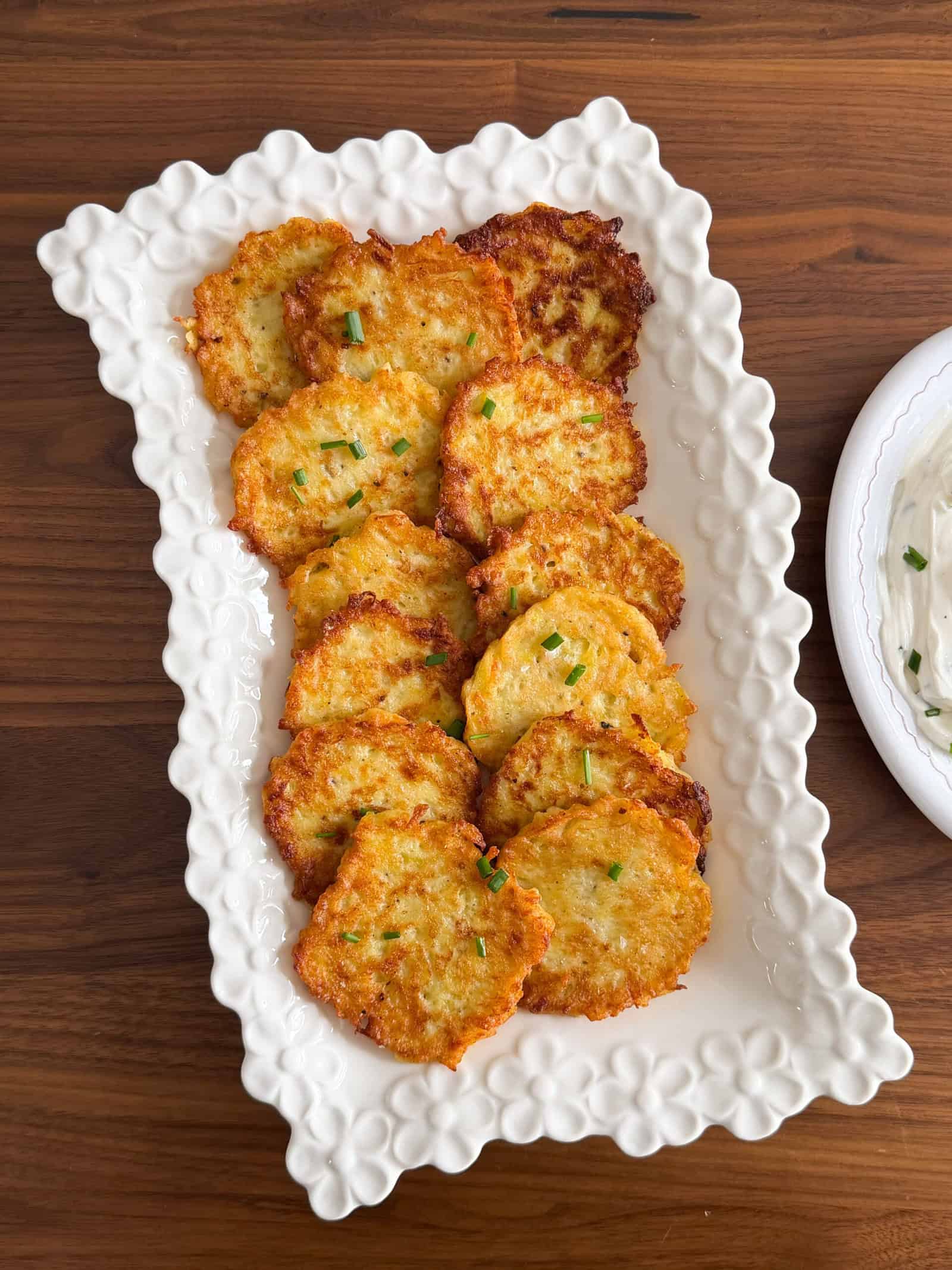A rectangular white platter holds a neat row of golden, crispy German Potato Pancakes, garnished with chopped green herbs, on a wooden table. A dish of white dipping sauce is partially visible on the right.