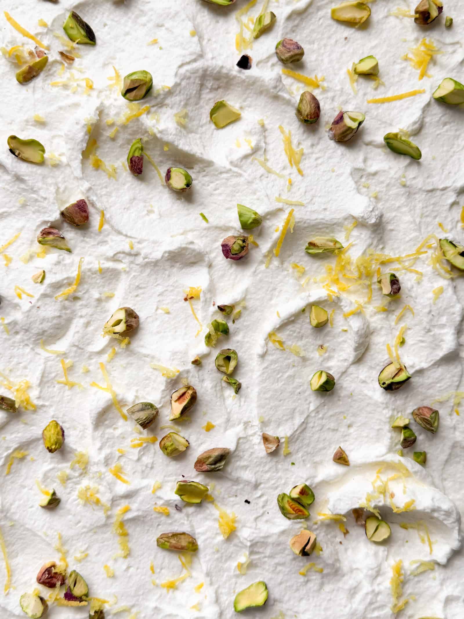 Whipped cream spread with a textured surface, topped with chopped pistachios and thin strands of lemon zest—an elegant finish perfect for any pistachio cheesecake recipe.