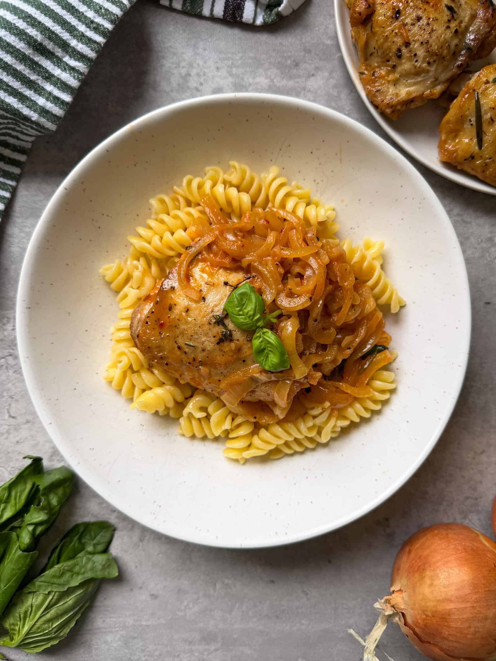 A white plate with rotini pasta topped with braised chicken and onions, caramelized to perfection, and fresh basil leaves, set on a light gray surface with fresh basil, an onion, and a striped towel nearby—a delicious choice for healthy chicken dinners.