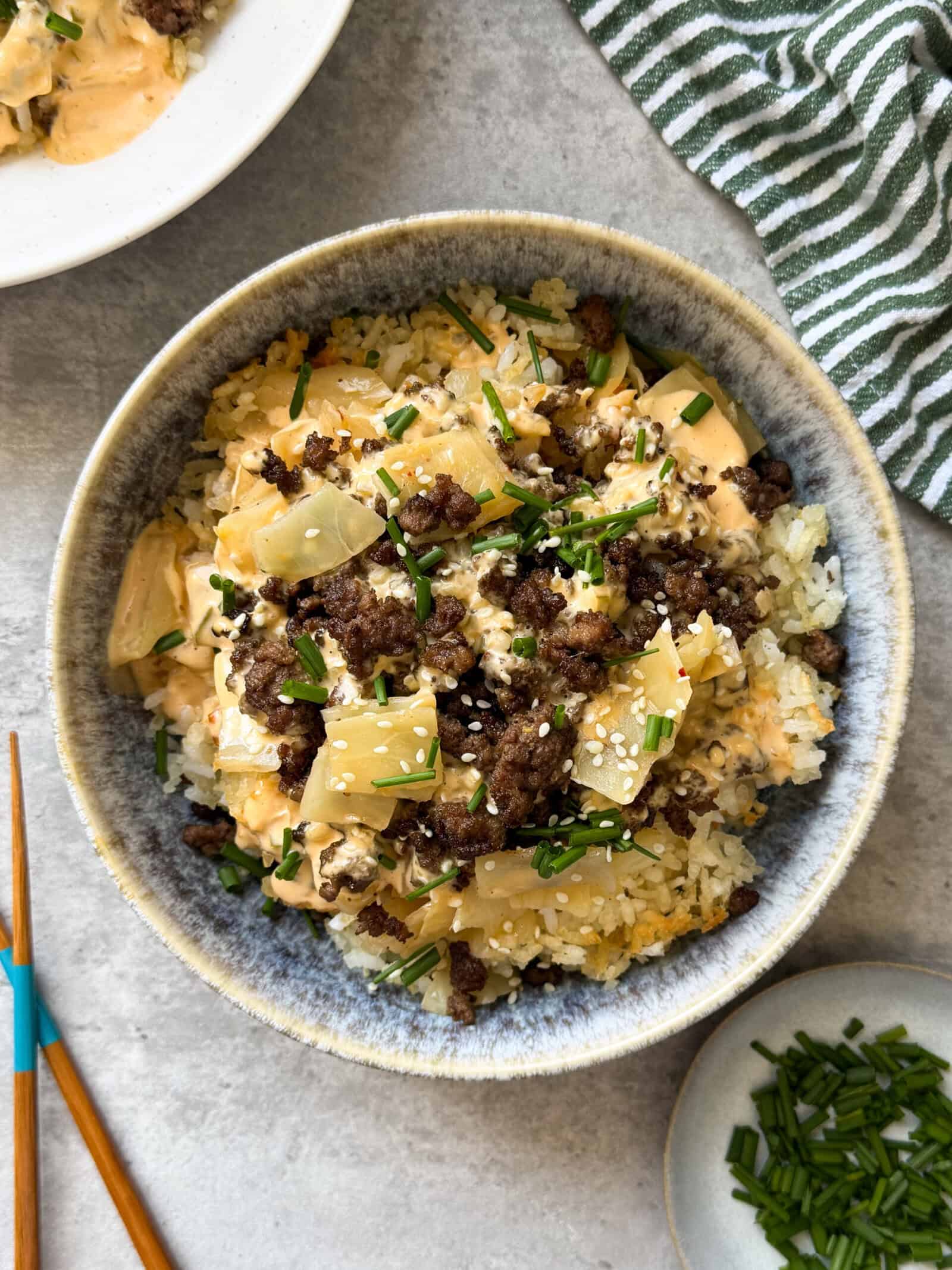 A bowl of rice topped with ground beef, chopped onions, creamy sauce, sesame seeds, and chives—like a Big Mac salad in a bowl. With chopsticks and a green-striped napkin nearby, it's perfect for fans of big mac cabbage and crispy rice bowls.