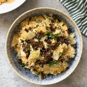 A bowl of rice topped with ground beef, chopped chives, sesame seeds, diced yellow vegetables, and a creamy sauce brings Big Mac salad vibes—perfect if you love Big Mac cabbage and crispy rice bowls. Set on a light gray surface with a green striped cloth nearby.