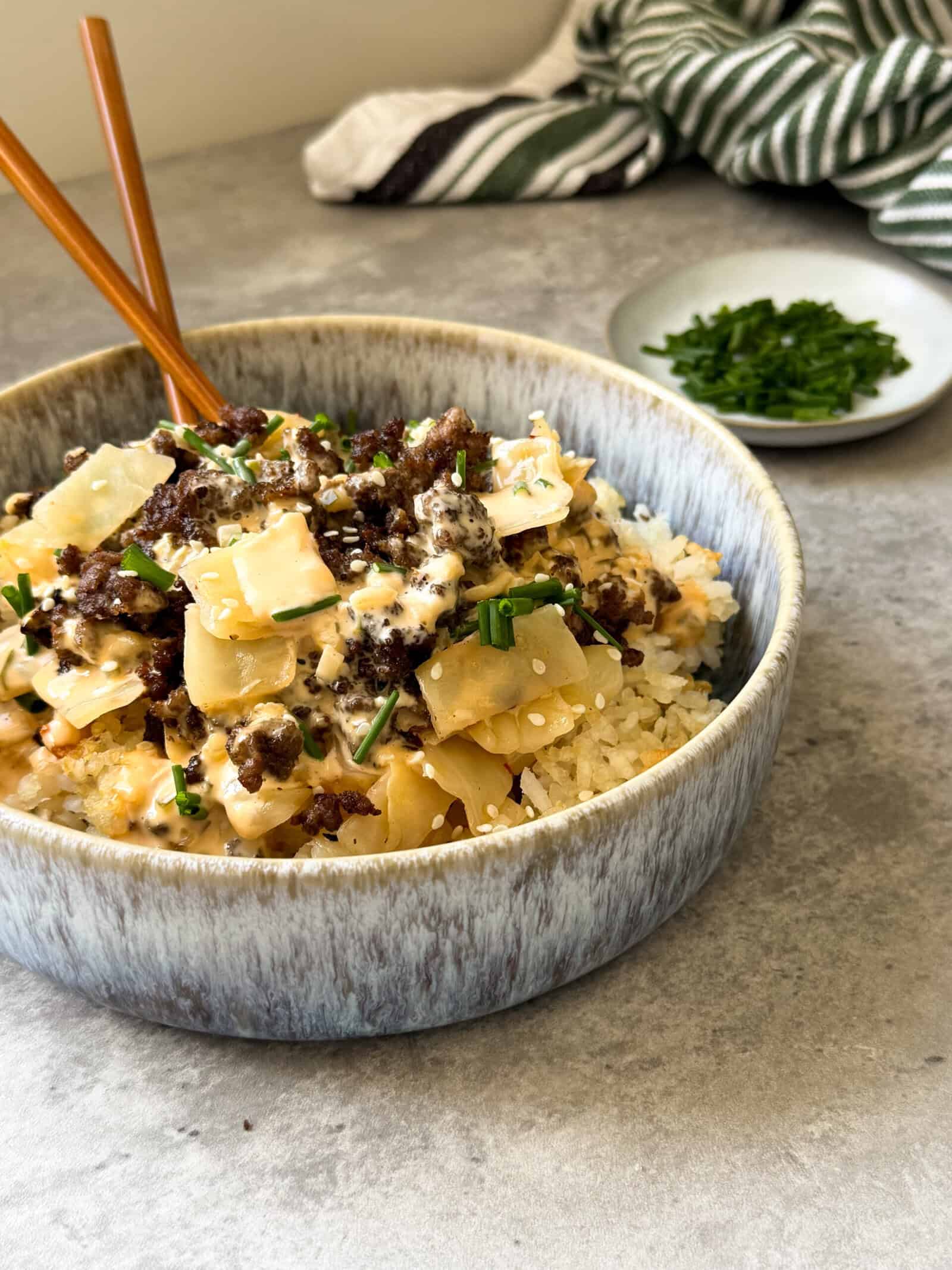A ceramic bowl filled with rice, ground beef, chopped onions, sauce, and a garnish of fresh chopped chives—like a big mac cabbage and crispy rice bowl. Wooden chopsticks rest inside; extra chives sit on a plate in the background on a gray surface.