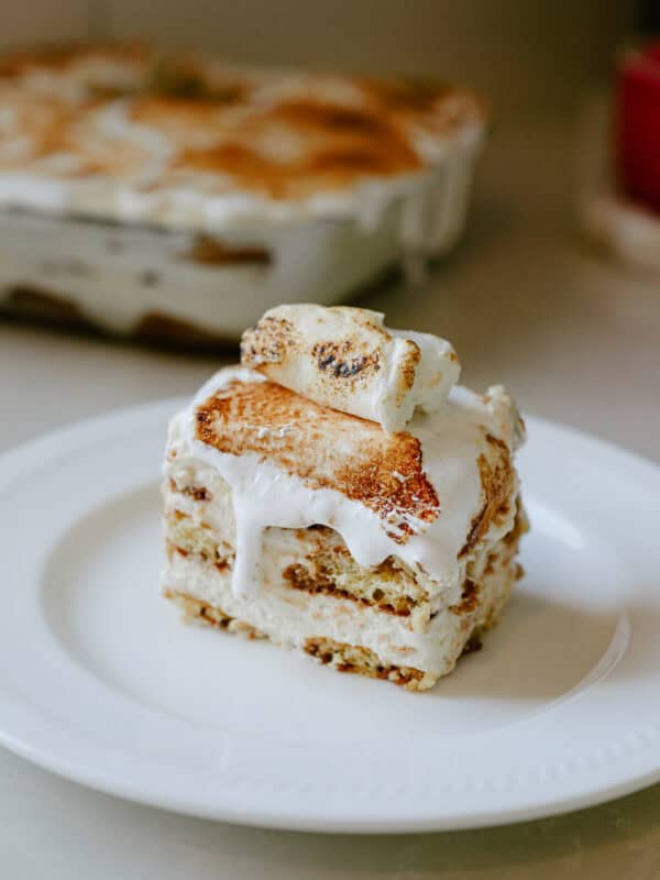A slice of creamy dessert with layers of cake and frosting is served on a white plate, with another dish of the same dessert blurred in the background.