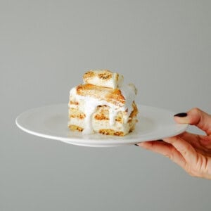A hand with black nail polish holds a white plate with a slice of layered cake topped with melting white icing against a plain, light gray background.