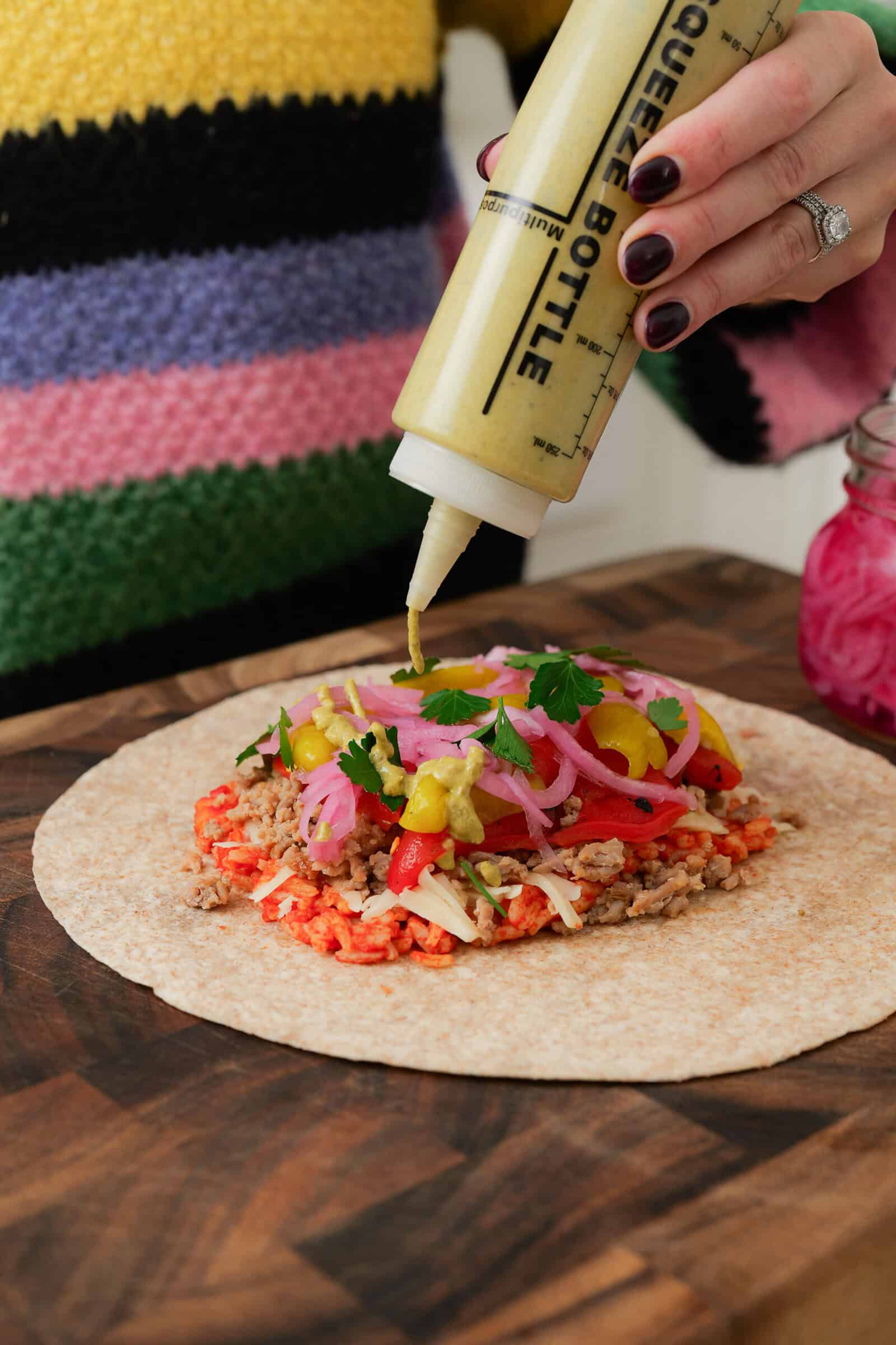 A person in a colorful sweater squeezes sauce from a bottle onto a tortilla wrap filled with ground meat, pickled onions, peppers, and fresh herbs on a wooden surface.