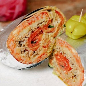 A close-up of a burrito cut in half, wrapped partly in foil, showing layers of ground meat, rice, cheese, tomatoes, and lettuce. A pepper and toothpick are on the side, with a jar of pickled onions in the background.