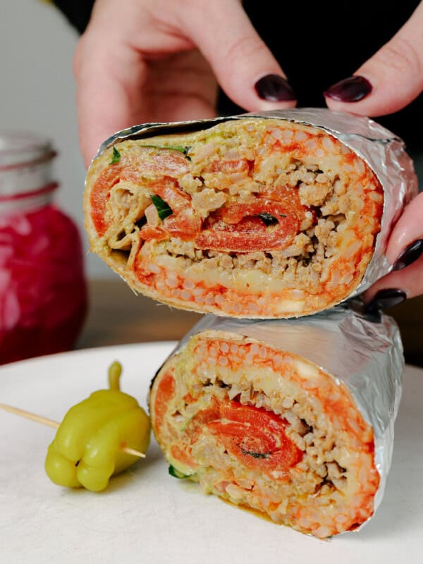 A person with dark nail polish holds a burrito cut in half, showing rice, ground meat, vegetables, and tomato inside. The burrito is wrapped in foil, and a pickled pepper sits on a white plate beside it.