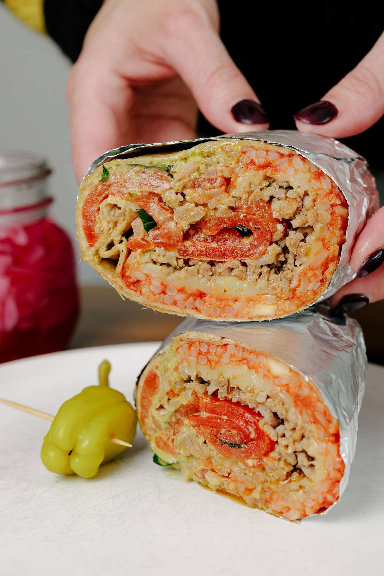 A person with dark nail polish holds a burrito cut in half, showing rice, ground meat, vegetables, and tomato inside. The burrito is wrapped in foil, and a pickled pepper sits on a white plate beside it.