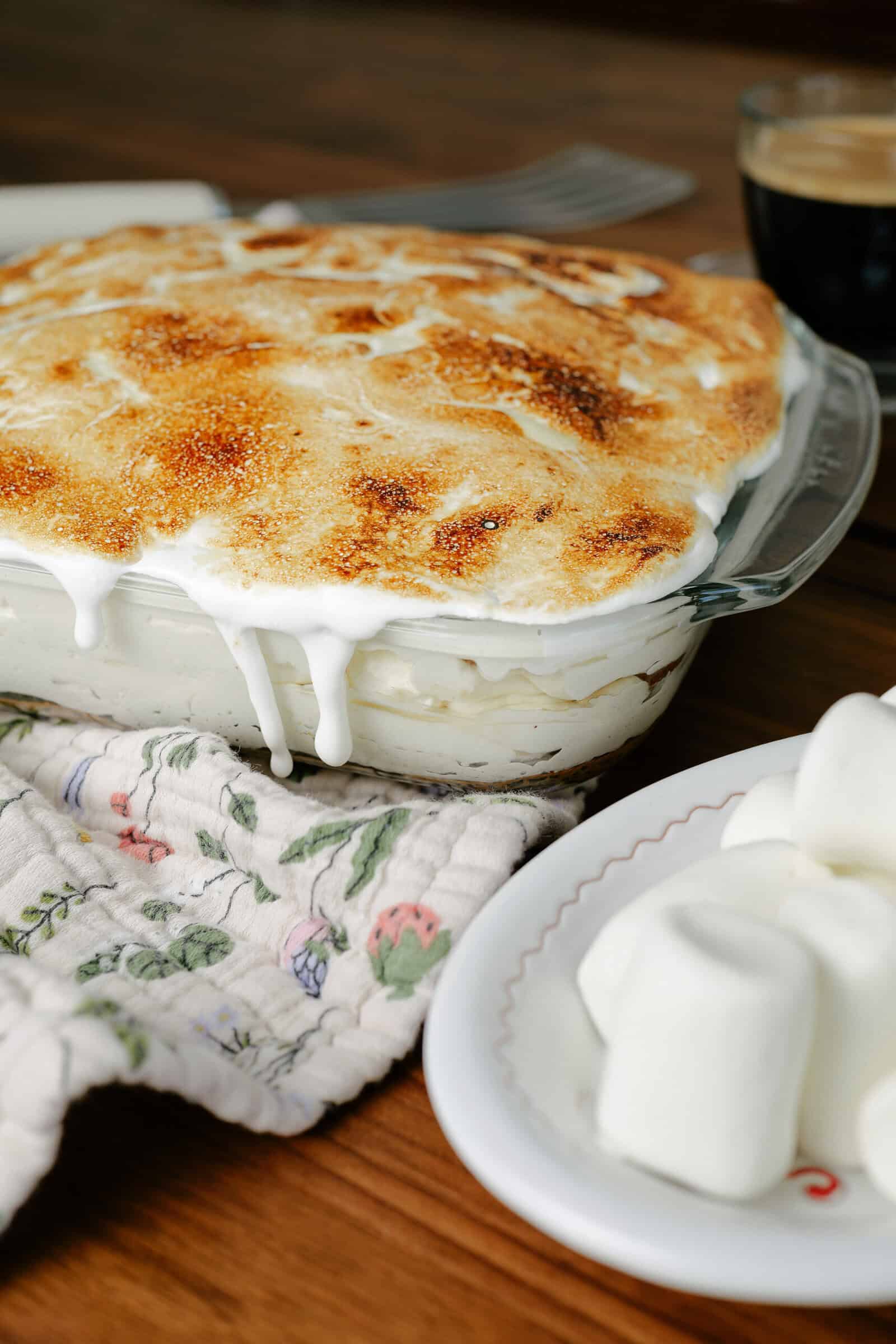 A baked dish with golden-brown toasted marshmallow topping drips over the edge, next to a floral oven mitt and a plate of marshmallows, with a cup of coffee in the background on a wooden table.
