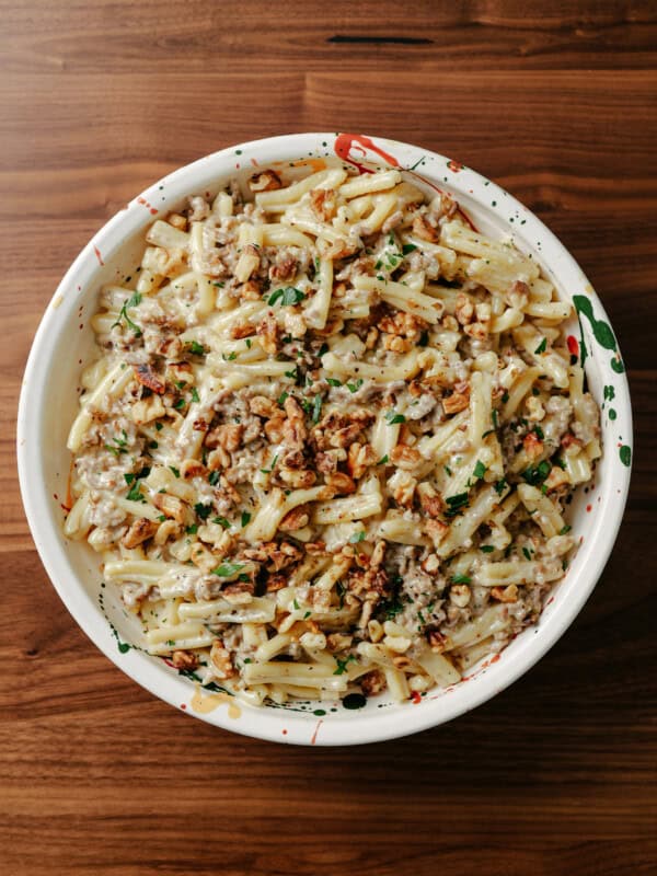 A bowl of creamy pasta mixed with ground meat and topped with chopped herbs and crushed nuts, placed on a wooden table.