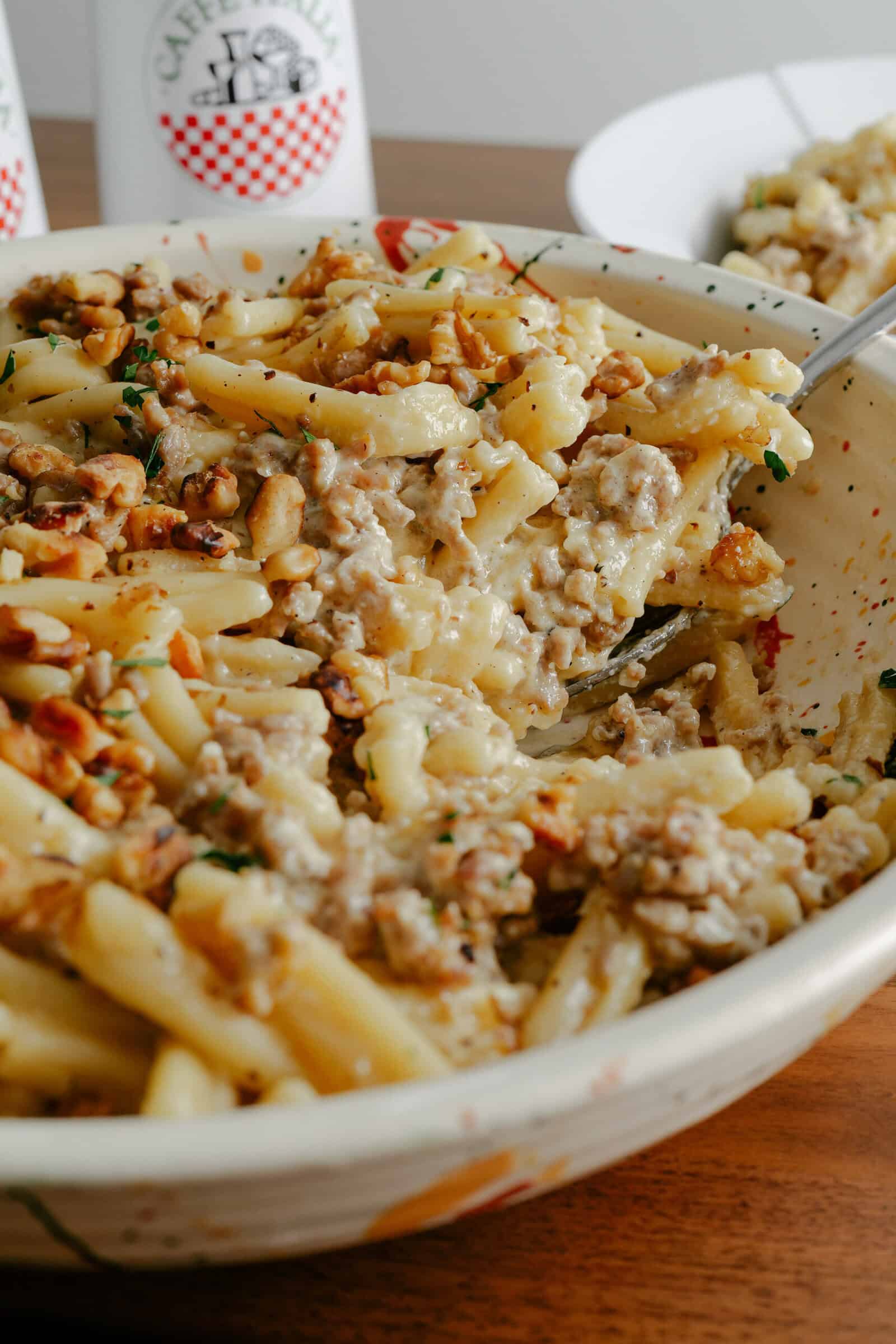 A close-up of a bowl of creamy pasta with ground sausage and chopped herbs, being stirred with a spoon. The dish is served on a wooden table with part of a white plate in the background.