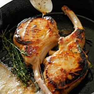 Two seared bone-in pork chops cooking in a cast iron skillet with fresh herbs and melted butter, while a spoon drizzles pan juices over the meat.