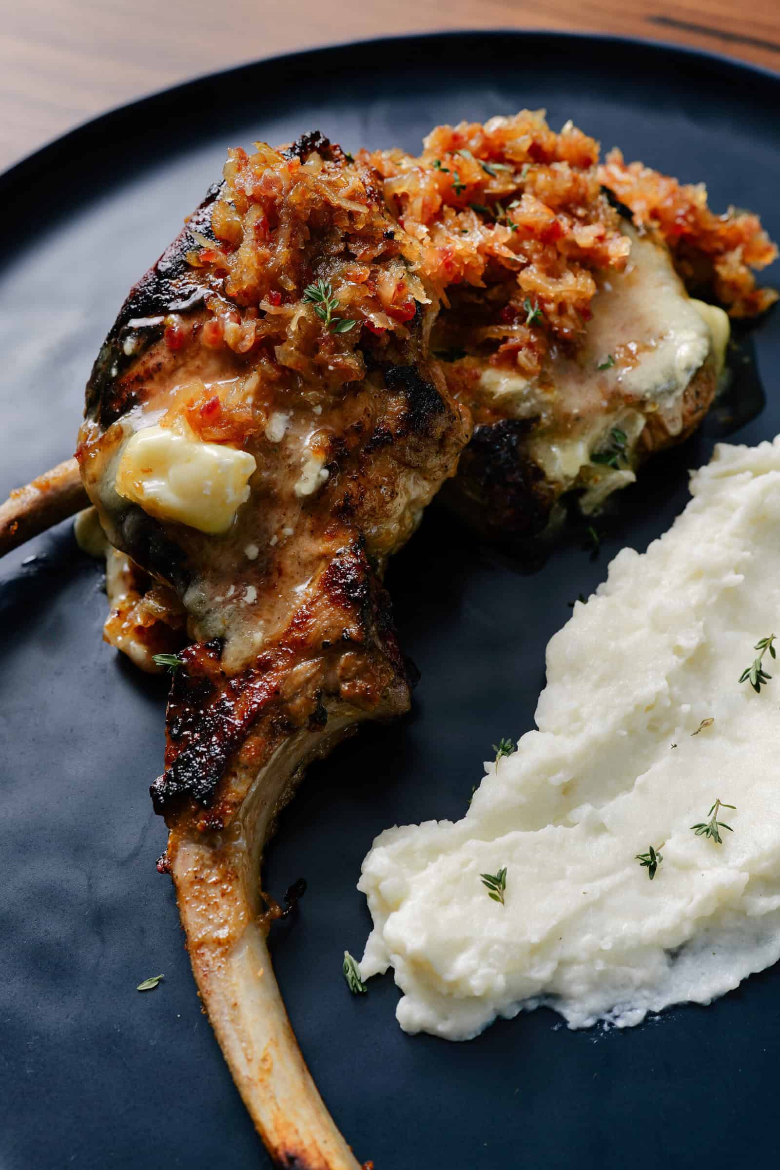 Two grilled lamb chops topped with a golden, chunky sauce, served next to a creamy scoop of mashed potatoes garnished with fresh herbs on a dark plate.