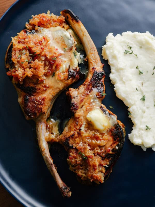 Two grilled lamb chops topped with a chunky vegetable relish, served next to a portion of creamy mashed potatoes garnished with fresh herbs on a dark plate.