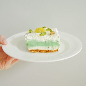 A hand holds a white plate with a slice of pistachio cheesecake featuring a green layer, crumbly base, whipped cream topping, and garnished with lemon slice and pistachios. The background is plain and light-colored.
