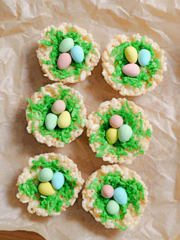 Seven Easter Rice Krispie treats shaped like nests, topped with green dyed shredded coconut and colorful candy eggs, are arranged on brown parchment paper—perfect festive rice krispie nests for your Easter treats spread.