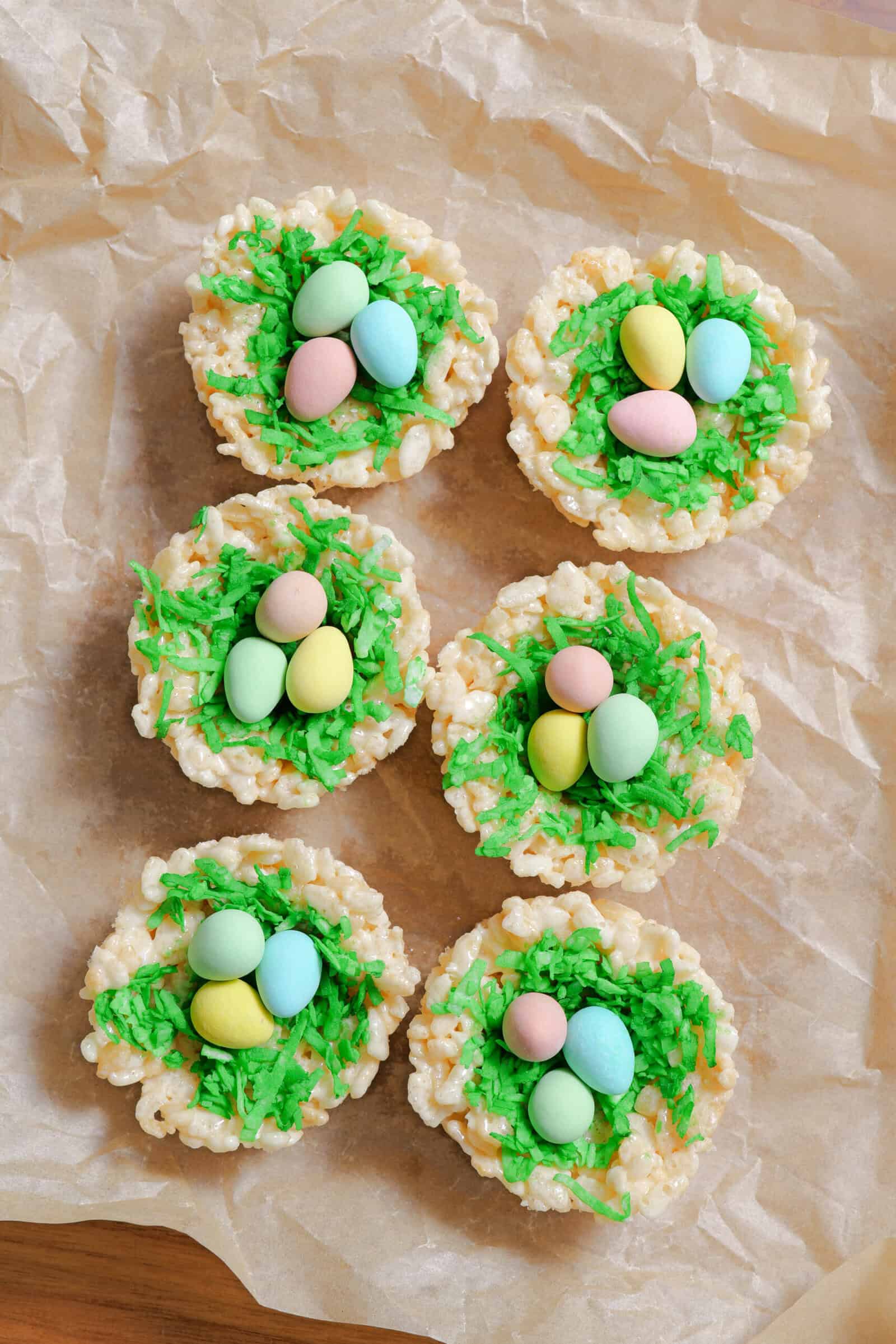 Seven Easter Rice Krispie treats shaped like nests, topped with green dyed shredded coconut and colorful candy eggs, are arranged on brown parchment paper—perfect festive rice krispie nests for your Easter treats spread.