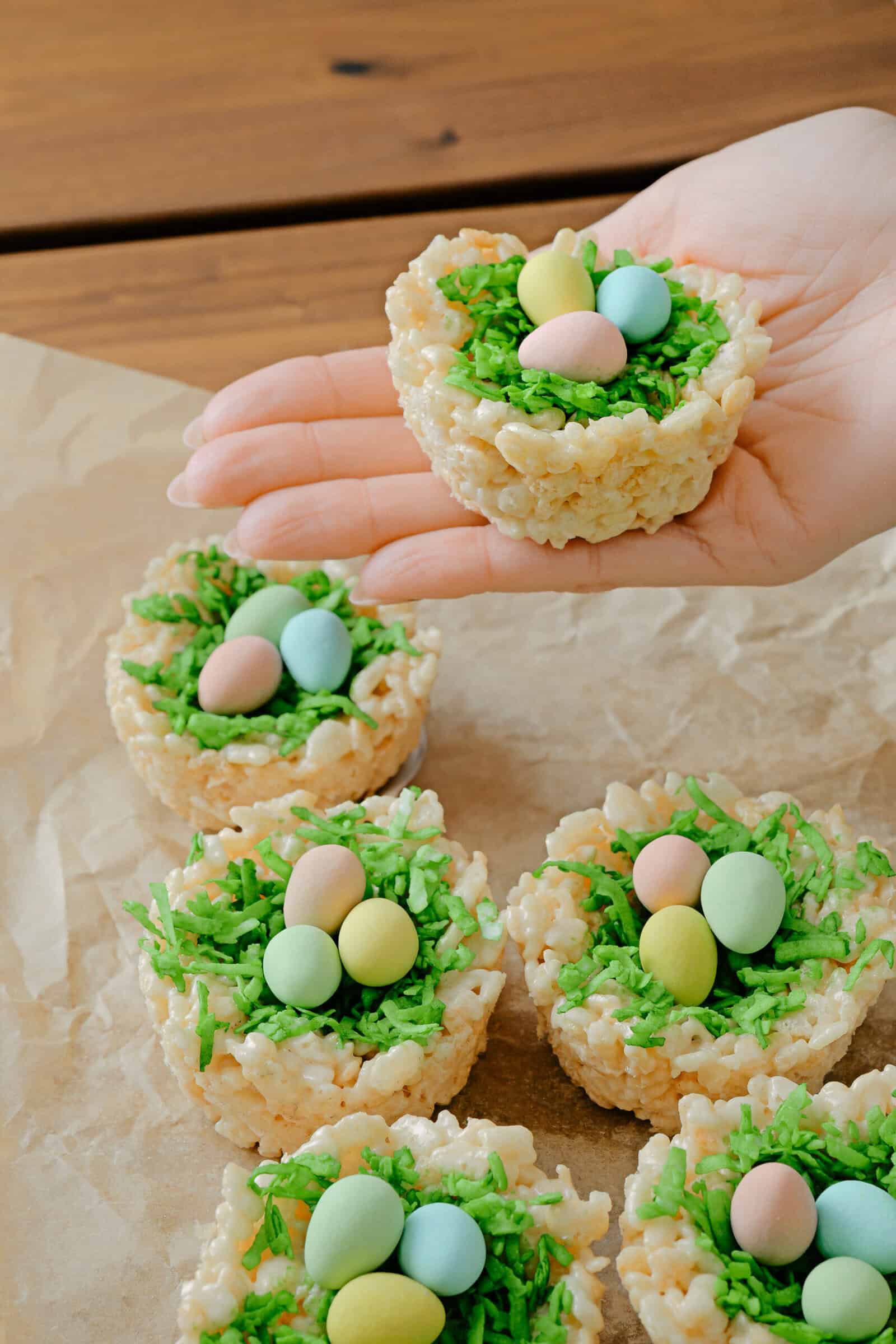 A hand holds a festive Rice Krispie nest, topped with green coconut “grass” and pastel candy eggs—perfect for Easter treats. More Rice Krispie nests rest on brown parchment paper below, ready to share.