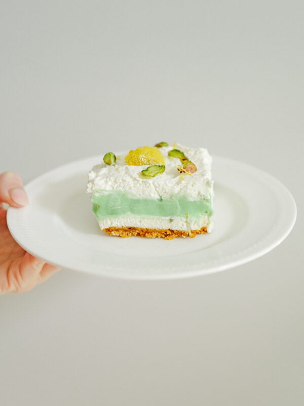 A hand holds a white plate with a slice of pistachio cheesecake featuring a golden crust, green filling, whipped topping, and garnished with pistachios and a lemon slice—perfect for anyone seeking an easy cheesecake recipe.