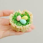 A hand holds a Rice Krispies treat shaped like a bird's nest, filled with green shredded coconut grass and three pastel-colored candy eggs—perfect for Easter treats or trying a fun rice krispie nests recipe.