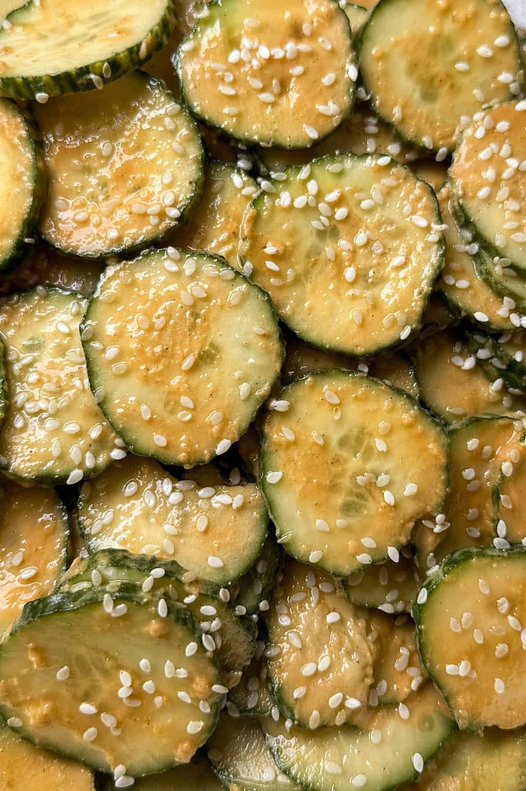 Close-up of a peanut butter cucumber salad: sliced cucumbers are coated in a creamy, orange-colored dressing and sprinkled with white sesame seeds.