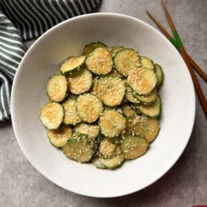 A white bowl filled with spicy peanut cucumber salad, topped with sesame seeds, sits on a gray surface, with wooden chopsticks and a green-striped towel beside it.