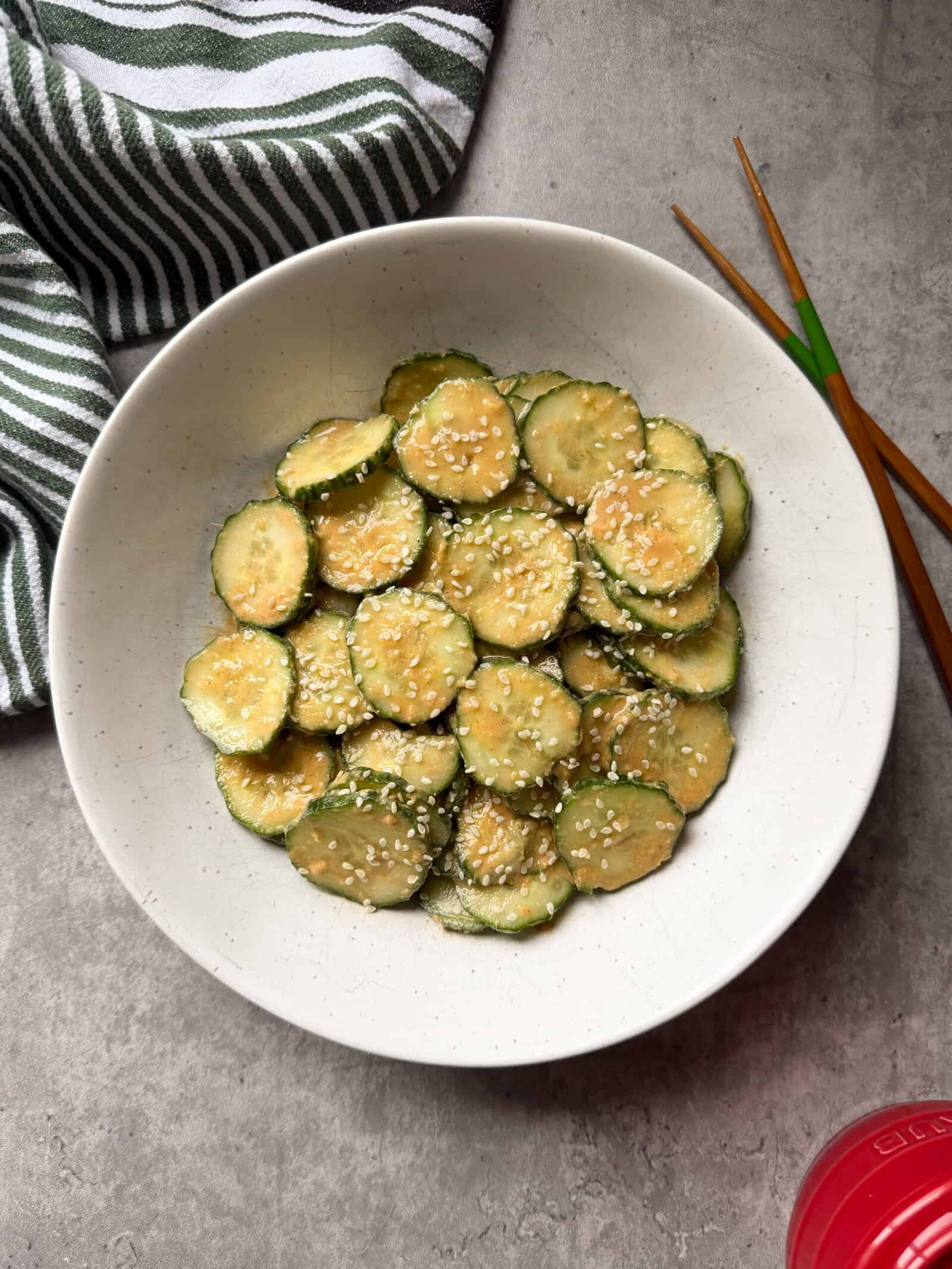 A white bowl filled with spicy peanut cucumber salad, topped with sesame seeds, sits on a gray surface, with wooden chopsticks and a green-striped towel beside it.