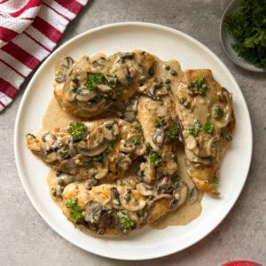 A white plate with Chicken Marsala&mdash;chicken breasts topped with creamy mushroom and caper sauce, garnished with parsley&mdash;sits on a gray surface beside a red-striped towel and a small dish of fresh parsley.