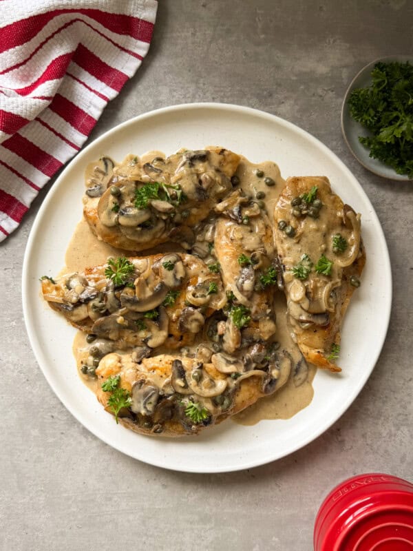 A white plate with Chicken Marsala—chicken breasts topped with creamy mushroom and caper sauce, garnished with parsley—sits on a gray surface beside a red-striped towel and a small dish of fresh parsley.