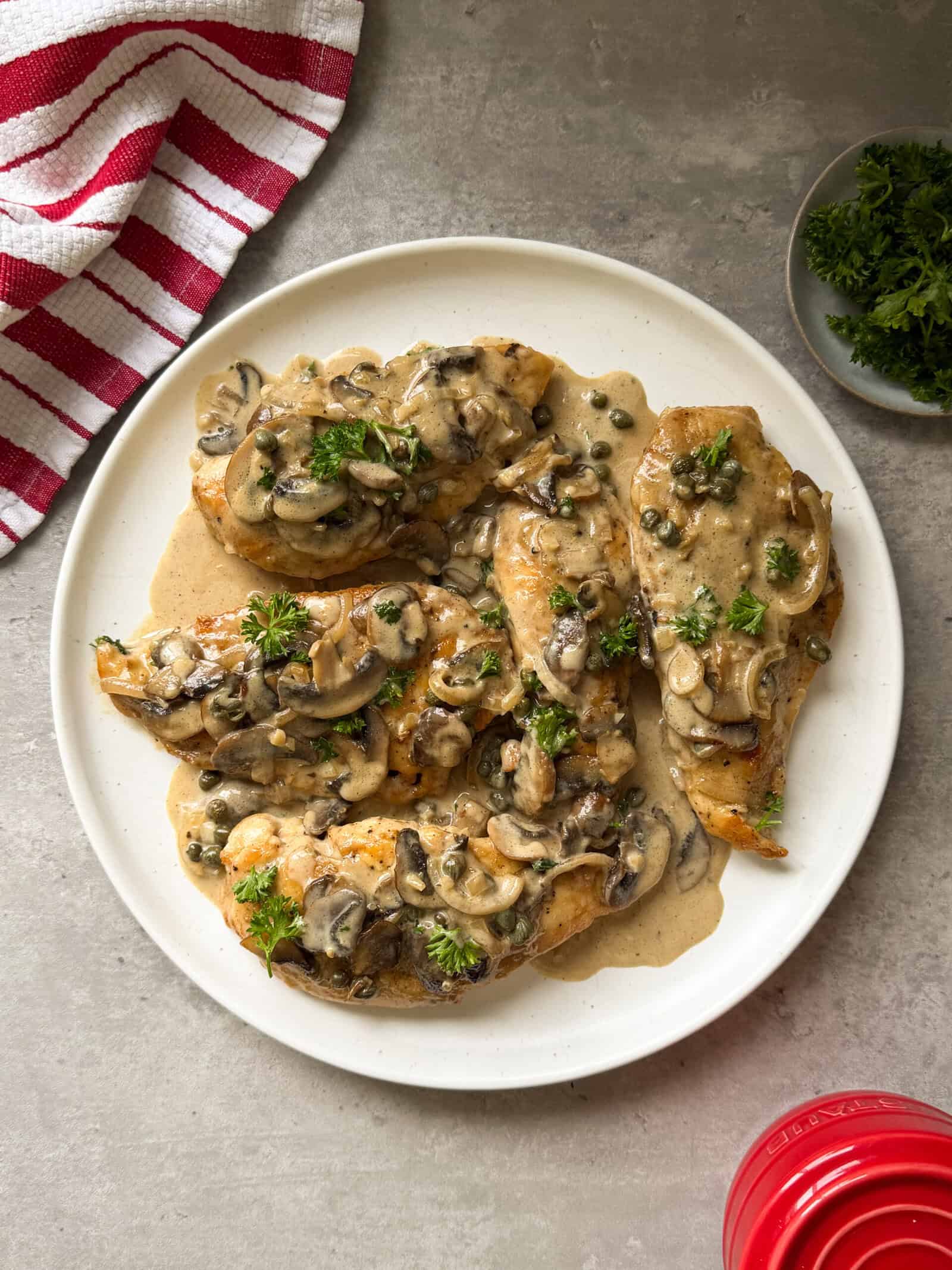 A white plate with Chicken Marsala&mdash;chicken breasts topped with creamy mushroom and caper sauce, garnished with parsley&mdash;sits on a gray surface beside a red-striped towel and a small dish of fresh parsley.