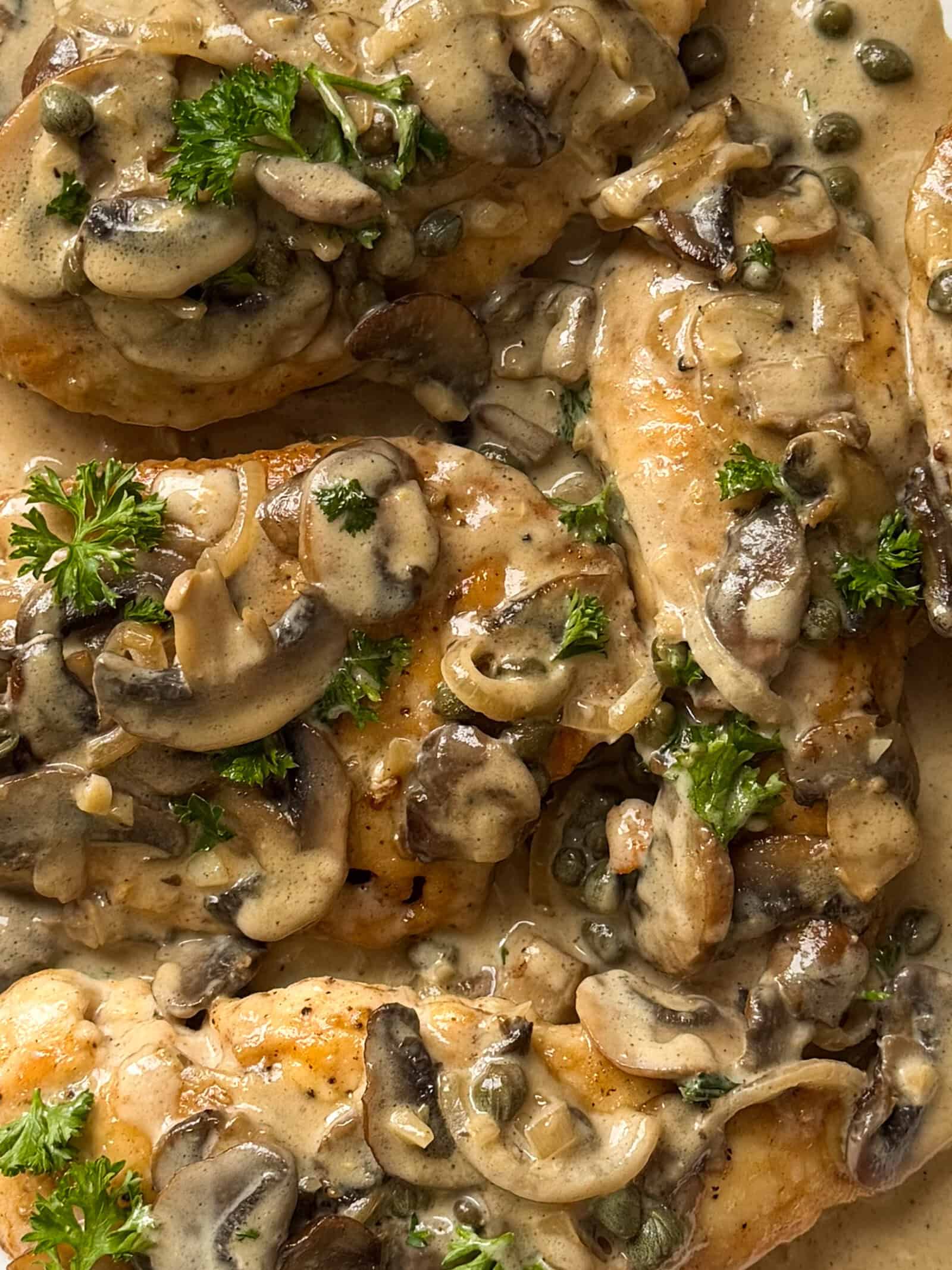 Close-up of creamy Chicken Marsala breast topped with saut&eacute;ed mushrooms, capers, and fresh parsley, all coated in a rich, light-brown sauce.