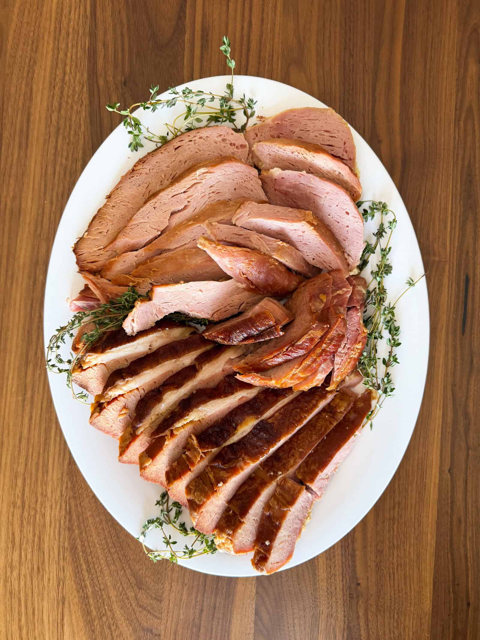 Sliced glazed ham arranged on a white oval platter, garnished with fresh thyme sprigs, set on a wooden table—perfect for showcasing your favorite crockpot ham recipe.