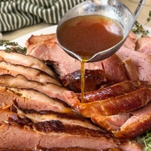A ladle pours brown glaze over slices of Crock-Pot Ham arranged on a plate, with fresh herbs and a green striped towel in the background.