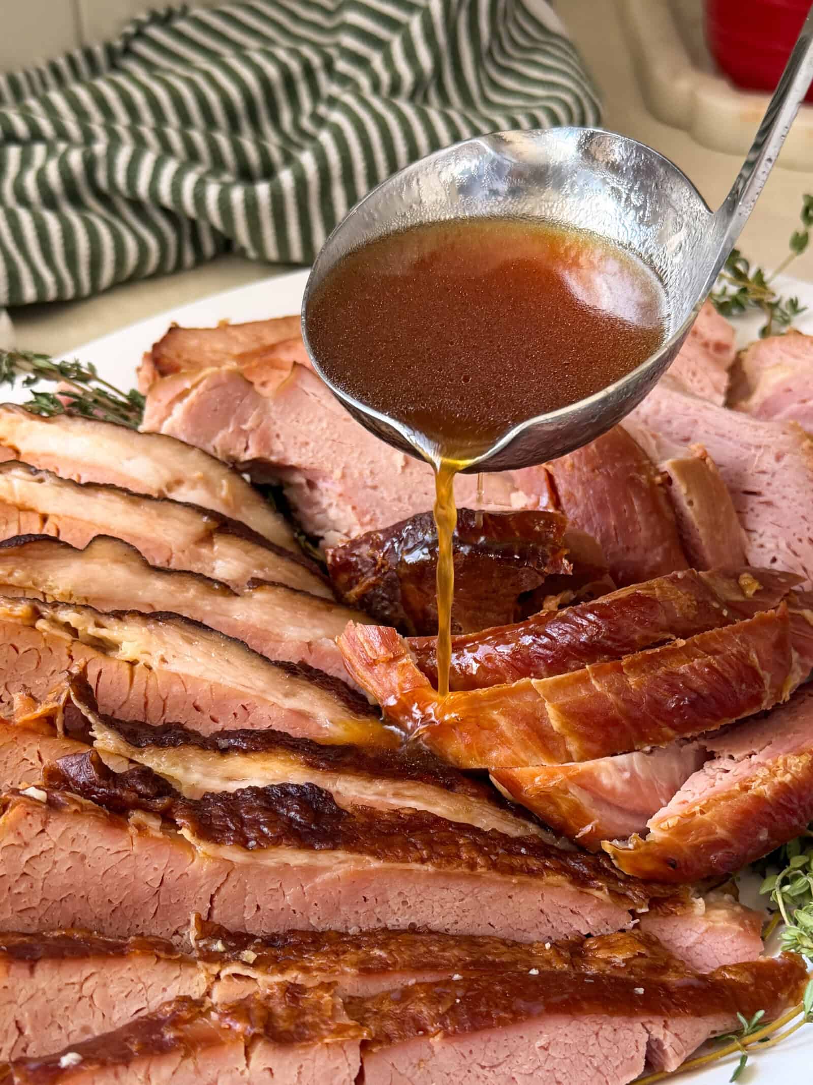 A ladle pours brown glaze over slices of Crock-Pot Ham arranged on a plate, with fresh herbs and a green striped towel in the background.