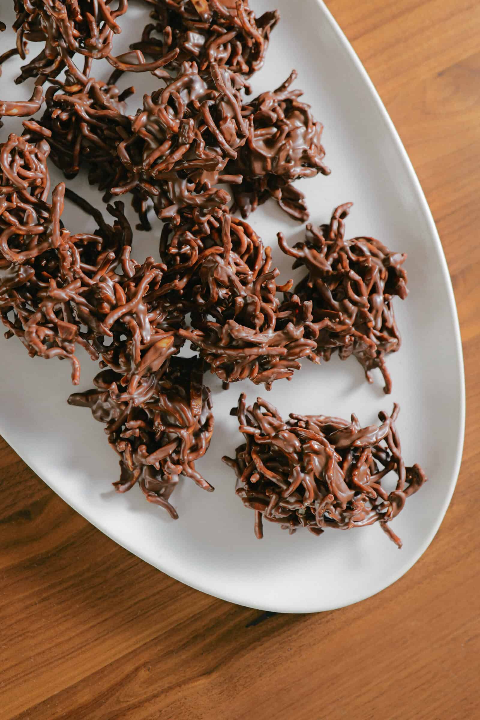 A white plate on a wooden surface holds clusters of chocolate-covered chow mein noodle cookies arranged in a casual pile—an irresistible treat for fans of easy no bake desserts and classic haystack recipes with chow mein noodles.