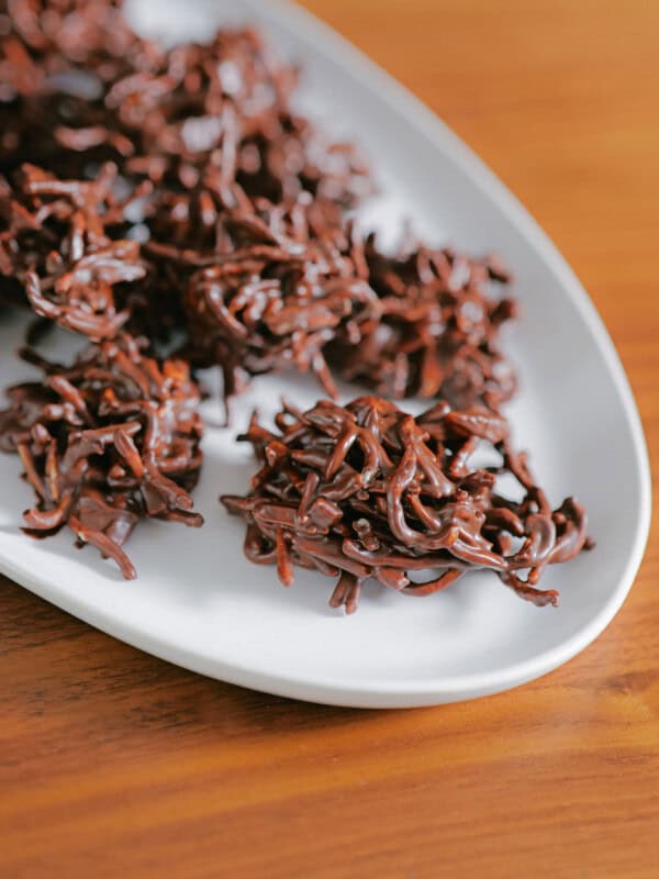 Chocolate haystack cookies arranged on a white plate, set on a wooden surface. These glossy chocolate chow mein clusters feature textured, noodle-like pieces and are perfect for those seeking easy no bake desserts.