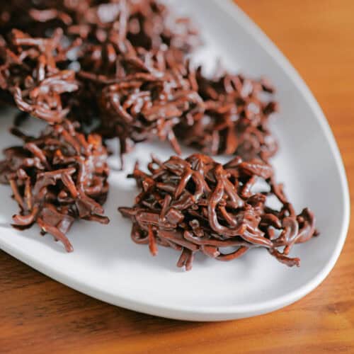 Clusters of chocolate-covered crispy chow mein noodles are arranged on a white oval plate, set on a wooden surface—an irresistible take on easy no bake desserts.