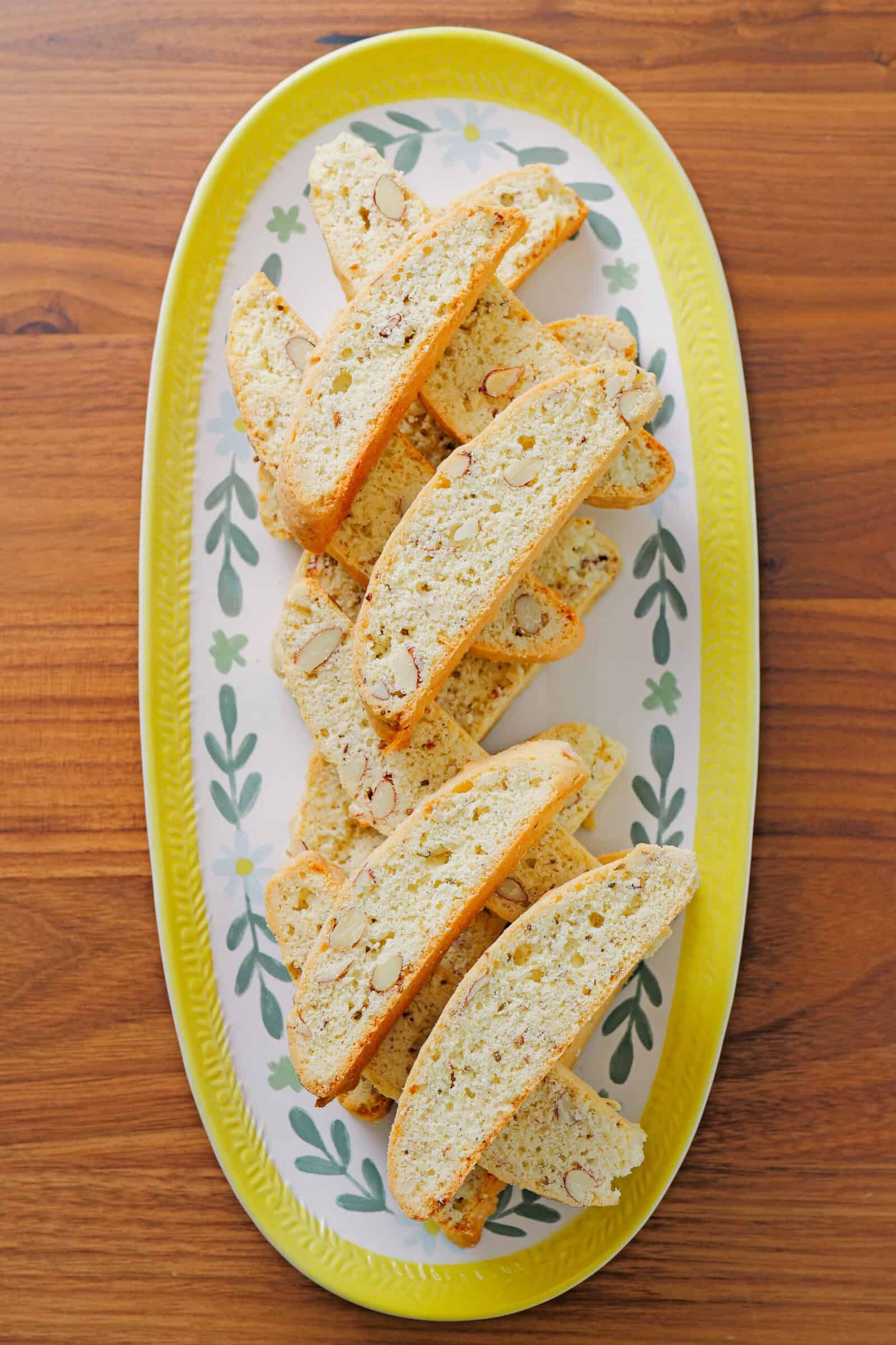 A decorative oval plate with a yellow rim holds several almond biscotti arranged in a neat pile on a wooden table.