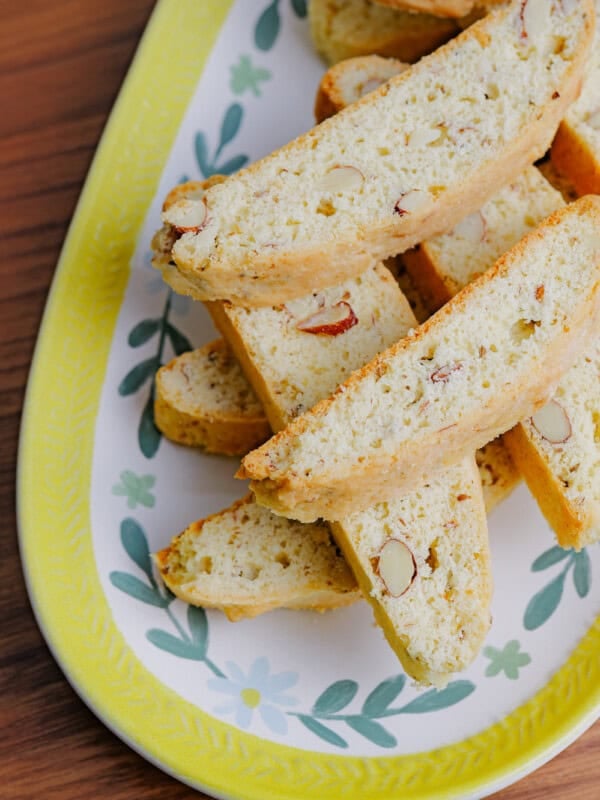 A close-up of several almond biscotti stacked on a decorative oval plate with a yellow and floral border, set on a wooden surface.