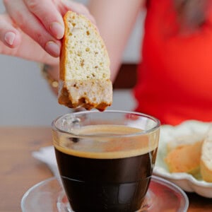 A person dips a slice of biscotti into a glass cup of black coffee on a wooden table. The person is wearing a bright orange shirt, and a plate with more biscotti is visible in the background.