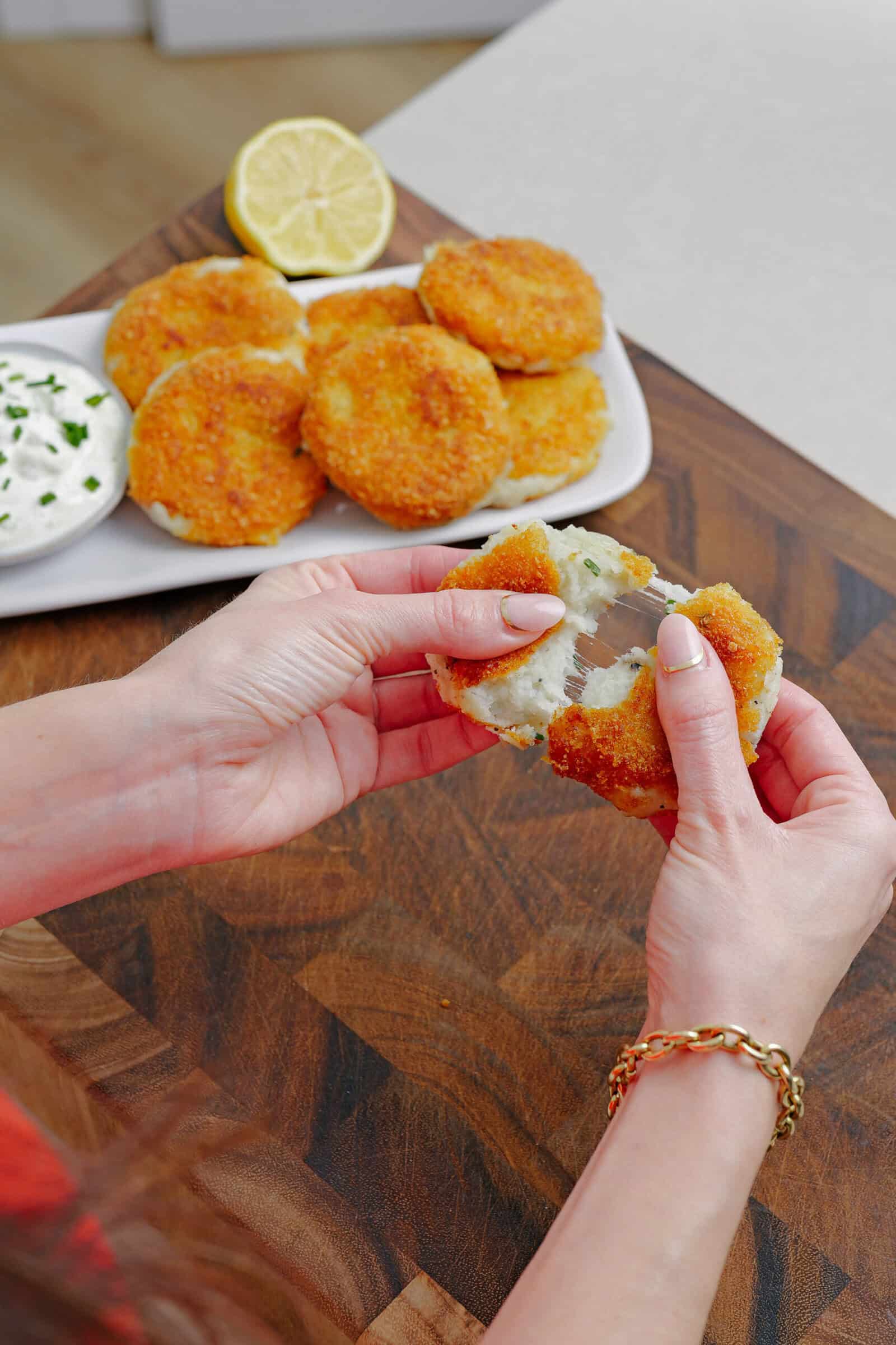 A person breaks open a golden, breaded patty, revealing a soft filling. In the background, more patties, a sliced lemon, and a bowl of dip with herbs sit on a white plate on a wooden surface.