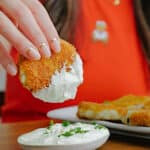 A person in a red shirt dips a breaded, fried food into a bowl of creamy white sauce garnished with chopped herbs. Another piece of the breaded food sits on a plate nearby.