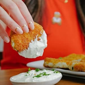A person in a red shirt dips a breaded, fried food into a bowl of creamy white sauce garnished with chopped herbs. Another piece of the breaded food sits on a plate nearby.