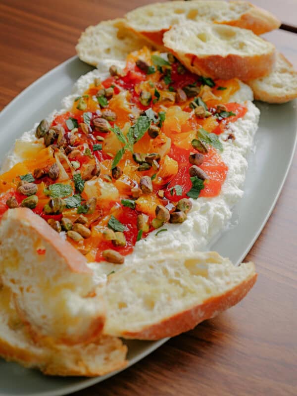 A platter of whipped ricotta topped with colorful roasted peppers, chopped herbs, and pistachios, served with slices of crusty bread on a wooden table.