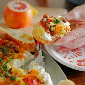 A hand holds a slice of bread topped with cheese, orange marmalade, pistachios, and herbs over a decorated plate. A partially sliced loaf and a halved orange are visible on the table.