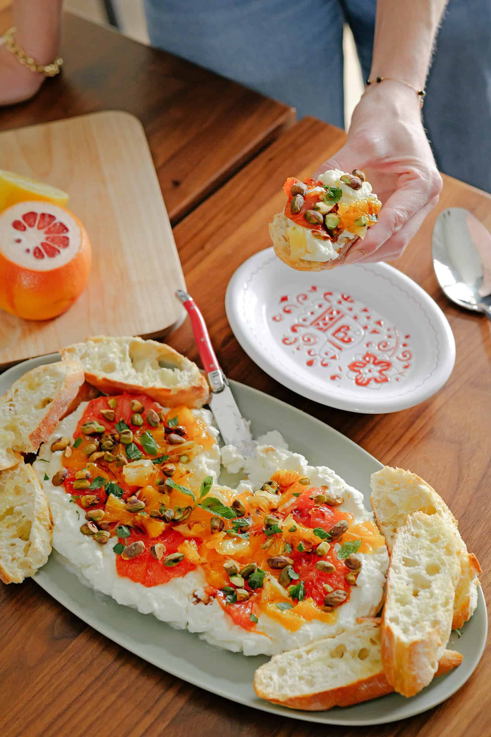 A person spreads colorful whipped cheese topped with herbs, seeds, and roasted peppers onto a slice of crusty bread. The dip is served on an oval platter with more bread slices, next to a plate and a halved grapefruit.