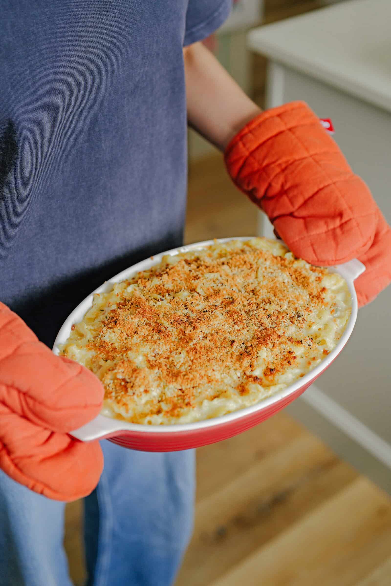 A person wearing orange oven mitts holds a baked dish of macaroni and cheese with a golden, crispy breadcrumb topping.