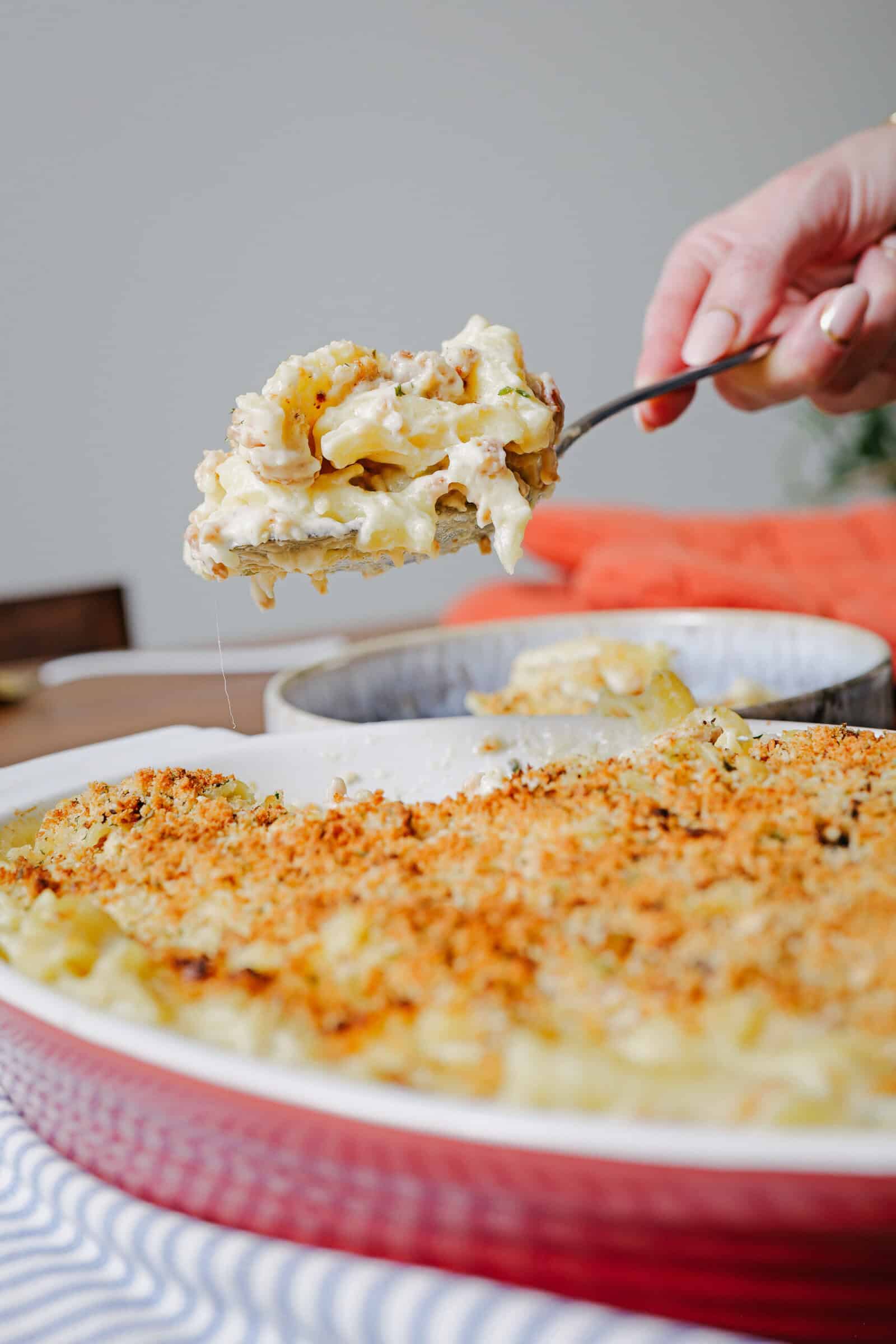 A hand holds a serving spoon scooping cheesy, baked macaroni and cheese with a golden breadcrumb topping from a red casserole dish.