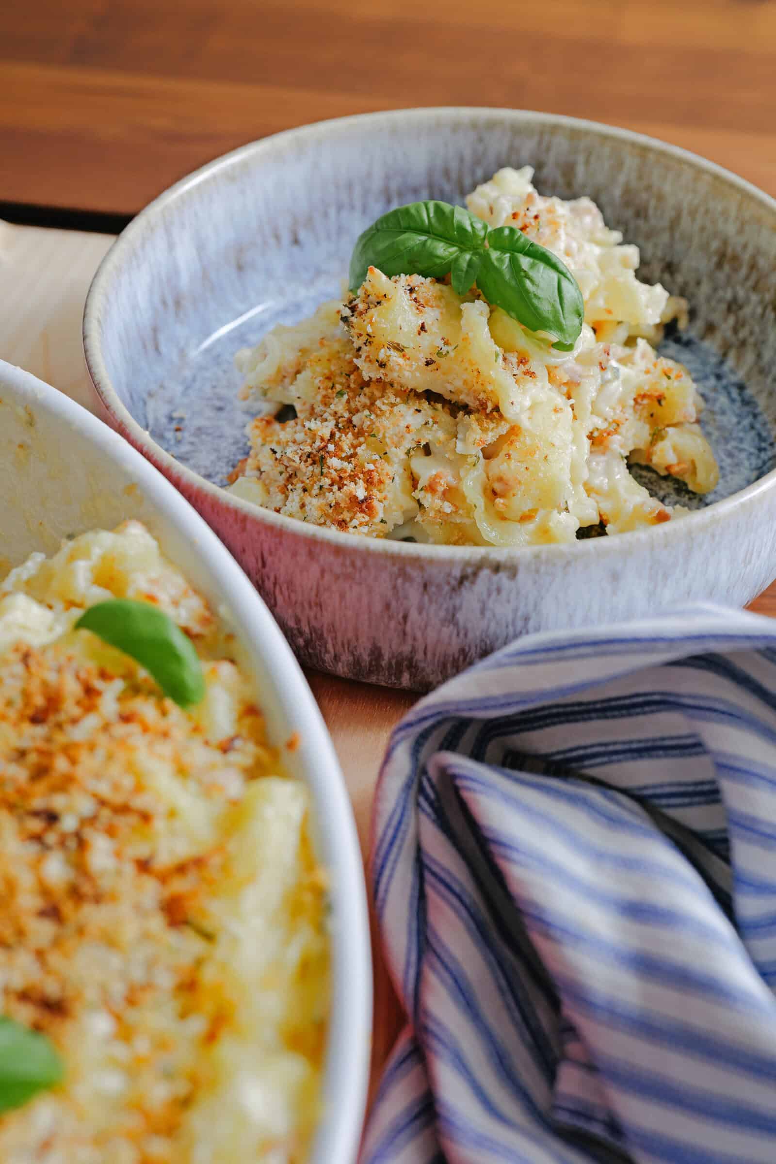 A bowl of baked macaroni and cheese topped with golden breadcrumbs and fresh basil, with a striped cloth beside it and a casserole dish visible in the foreground.